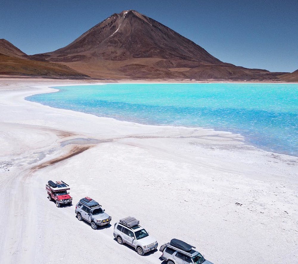 Vehículos estacionados al lado de laguna turquesa y volcán de fondo en Atacama